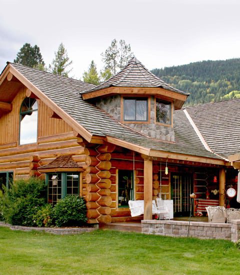 Exterior picture of a large log home with a lawn in the foreground and wooded hillside in the background.