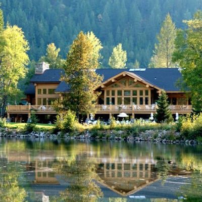 Large log and timber frame resort in a wooded area with a large pond in the foreground.