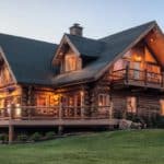 Chink style log home with exterior lights on and window illuminated. Fire in the firepit in the grassy yard.