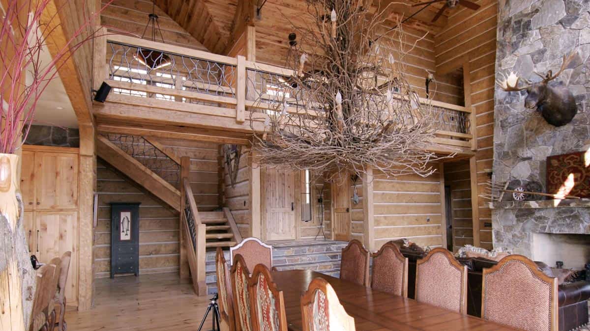 Dining room with a high ceiling, large rock fireplace featuring a mounted bull moose. Wooden railed staircase and second floor railings are visible..