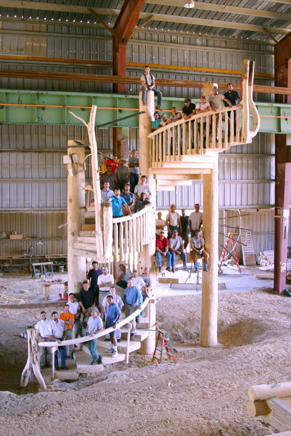 Custom built, three-story wooden spiral staircase in a large shop with 38 men standing on all different parts of the staircase.