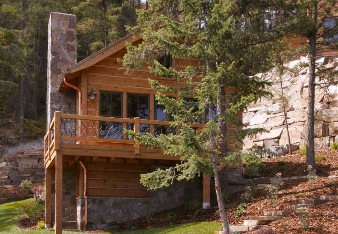 Two story log home nestled into the hillside with a stone wall behind it and trees in the foreground.
