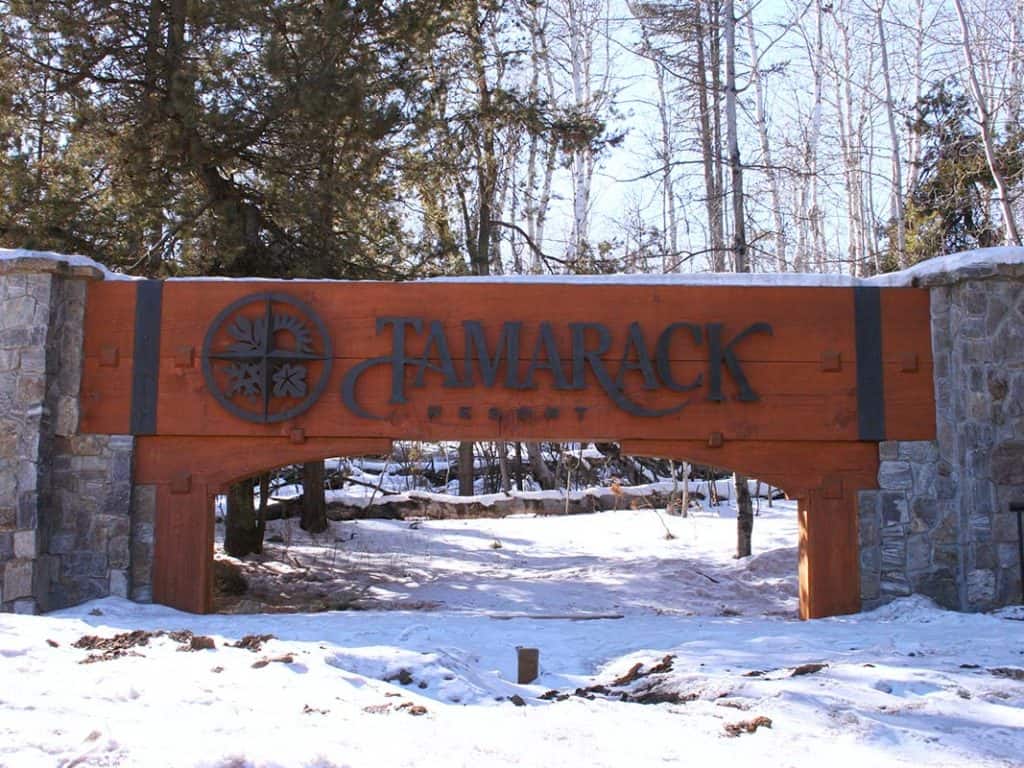 Large wooden sign for Tamarack Resort. Sign is between two stone supports, snow and trees all around the sign.