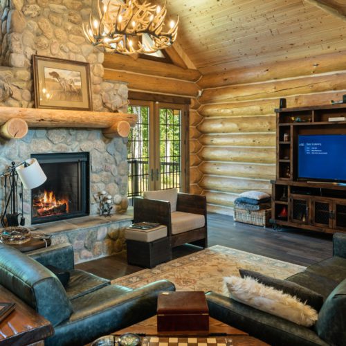 This modern log home is decorated with an antler chandelier, a stone fireplace, and gorgeous windows.